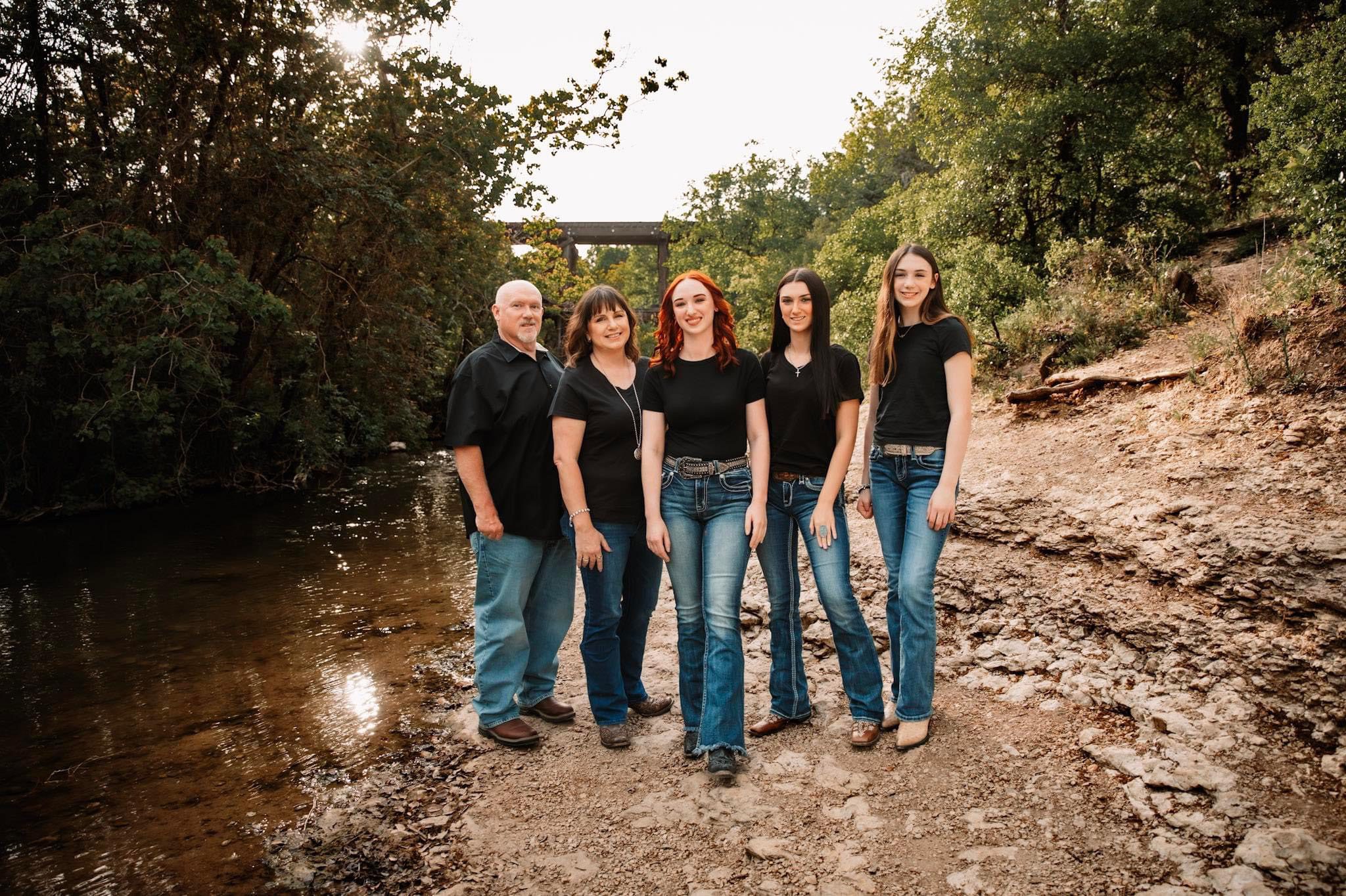 StainGuys-About-Team Five people stand on a rocky creek bank, wearing black shirts and blue jeans, with trees and a creek in the background.